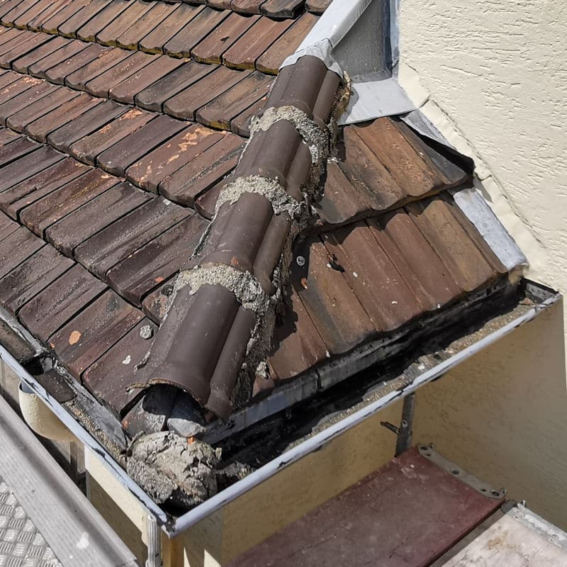 Ecke an einem Dach mit Firstdachziegeln, aus deren Ende ein großes Stück Mörtel herausgebrochen ist. Das muss dringend durch einen Dachdecker repariert werden.